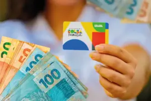 Mãos segurando cartão do Bolsa Família com logotipo colorido e notas de R$ 20 e R$ 100 em reais